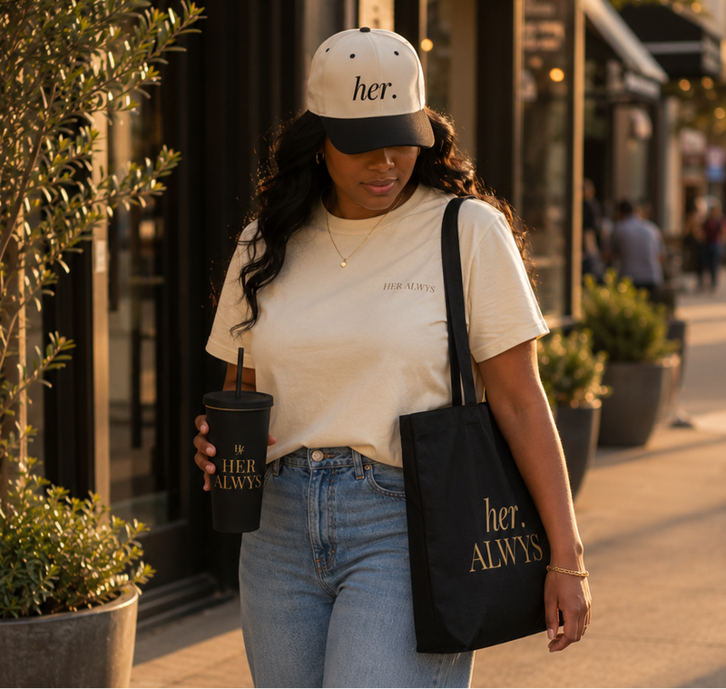 Woman walking outdoors holding a coffee cup and a black tote bag with 'her.' branding.